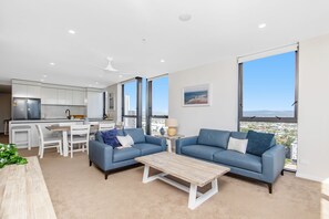 A modern living room with a sofa, coffee table, and a view of the city through large windows. - 2 Bedroom Broadbeach Stay – Walk to Beach (Broadbeach)