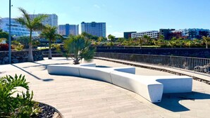 A modern outdoor area with a curved seating area, palm trees, and a view of high-rise buildings in the background. - Sol Paraiso 2 bedrooms apartment (Adeje)
