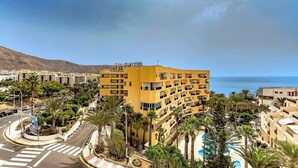 Un hotel amarillo de varios pisos con balcones, rodeado de palmeras y vista al mar.
