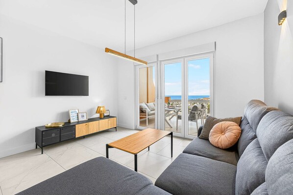 A modern living room with a sofa, a coffee table, a TV, and a sliding glass door leading to a balcony with a view.