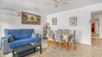 A living room with a blue sofa, a coffee table, a dining table with chairs, a ceiling fan, and wall-mounted air conditioning.
