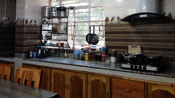 A kitchen with wooden cabinets, a gas stove, and a window with a view of greenery. - Bhagirath Homestay (Mirik)