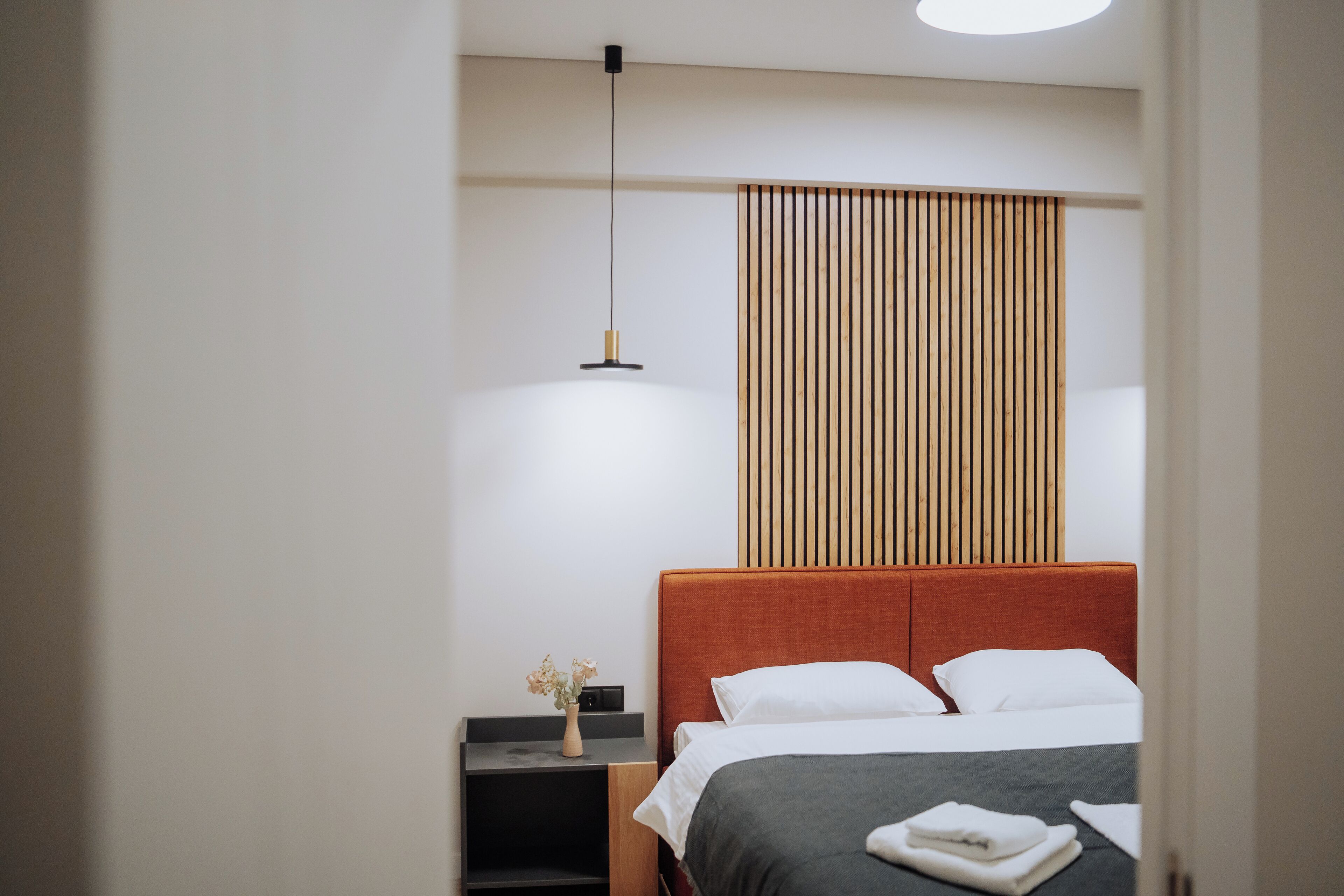 A modern hotel room with a large bed, a bedside table with a vase of flowers, and a wooden slat wall panel.