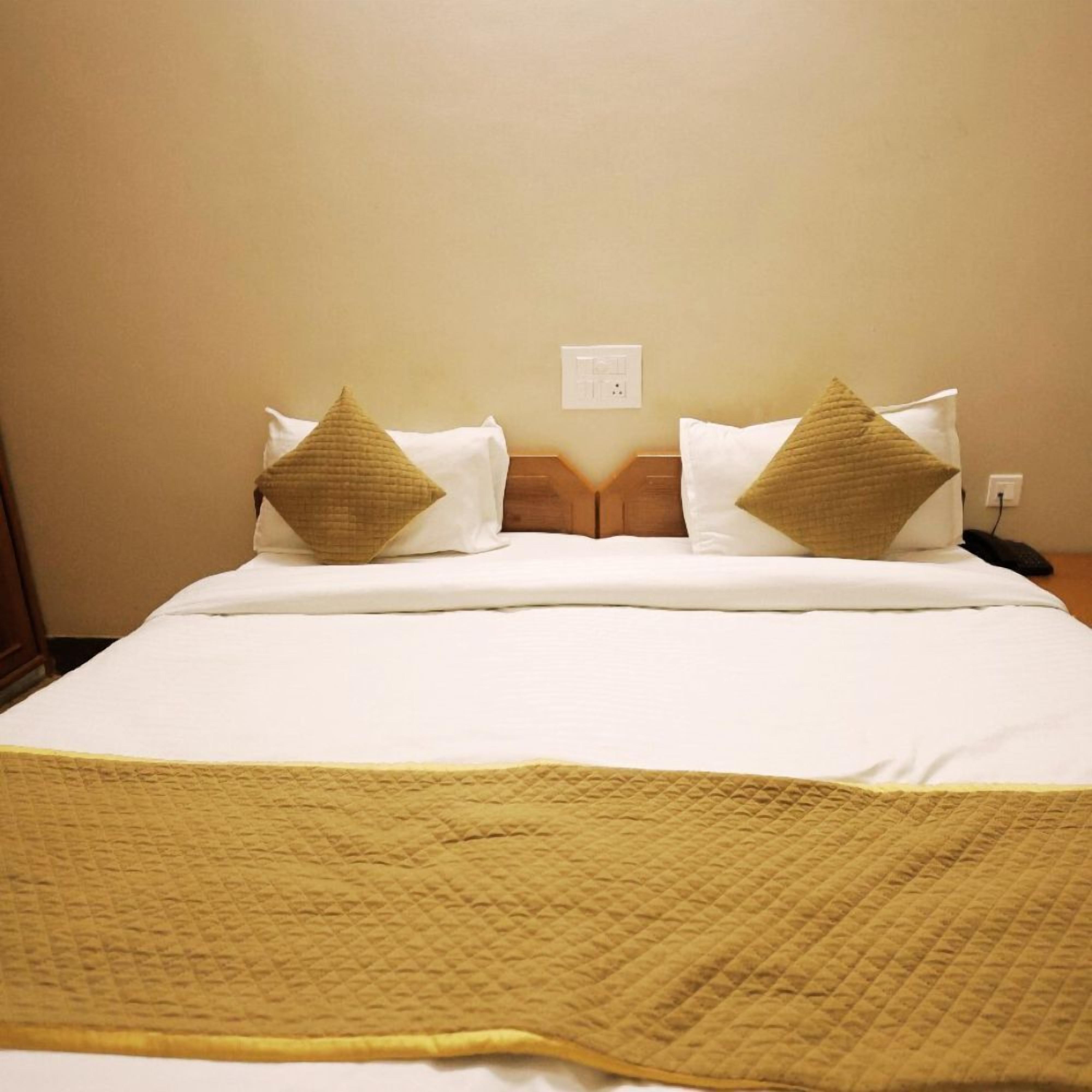 A neatly made bed with a white duvet and two beige pillows, a wooden headboard, and a power outlet on the wall.