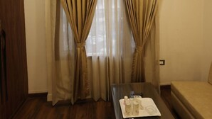 Una habitación de hotel con un armario de madera, una cama, una mesita con artículos de aseo y cortinas color beige.