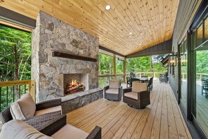 A covered deck with a stone fireplace, wicker furniture, and a view of the surrounding forest. - Sweet Bearadise-Eagles Nest - dogs ok, outdoor fireplace (Banner Elk)