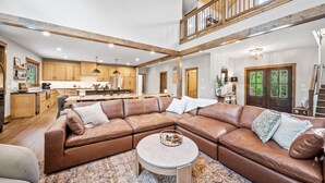 A spacious living area with a large brown leather sectional sofa, a round coffee table, and a high ceiling with exposed wooden beams.