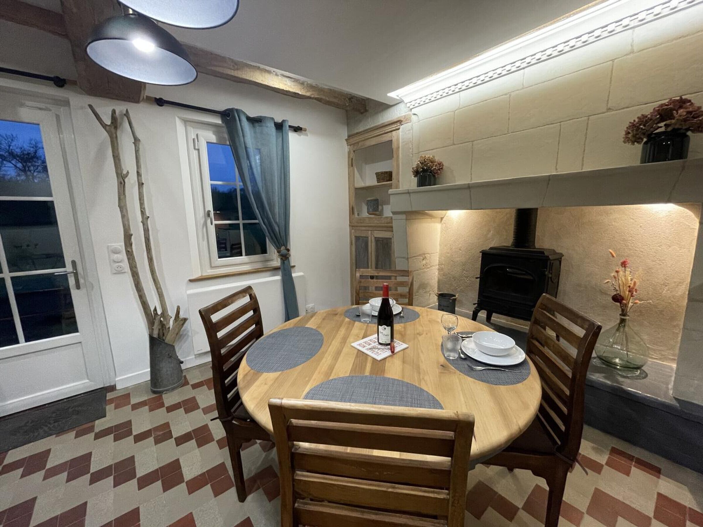 A dining area with a round wooden table, four chairs, a bottle of wine, and a vase with dried flowers. There is a fireplace, a window with curtains, and a checkered floor.