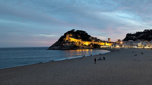 A beach with a historic castle on a hill, calm sea, and a few people on the sand.