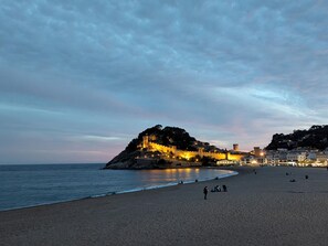 Una playa con un castillo histórico en una colina, mar tranquilo y algunas personas en la arena.