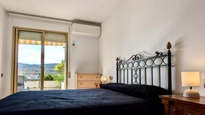 A bedroom with a bed, a wooden dresser, a lamp, and a view of a cityscape through a sliding door.