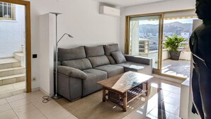 A modern living room with a grey sofa, a wooden coffee table, and a floor lamp. A large window offers a view of the cityscape.