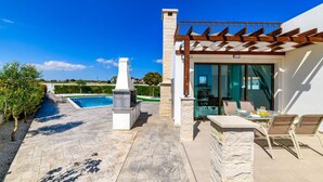 Une terrasse extérieure moderne, dotée d’un barbecue en pierre, d’une porte vitrée donnant sur un espace salon abrité et offrant une vue sur une piscine.