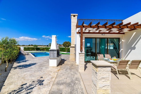 A modern outdoor patio with a stone barbecue, a glass door leading to a covered seating area, and a view of a swimming pool. - Kymma Seaview 24 in Ayia Napa (Sotira)