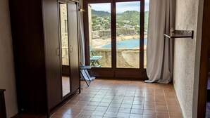 A room with a bed, a wardrobe, a window with a view of a beach and buildings, and a tiled floor.