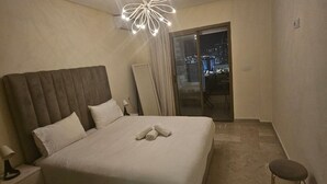 A hotel room with a large bed, a nightstand, a lamp, a chair, and a view of the city at night through a sliding door.