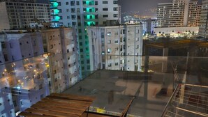 A rooftop terrace with a wooden table and metal railing, overlooking a cityscape at night.