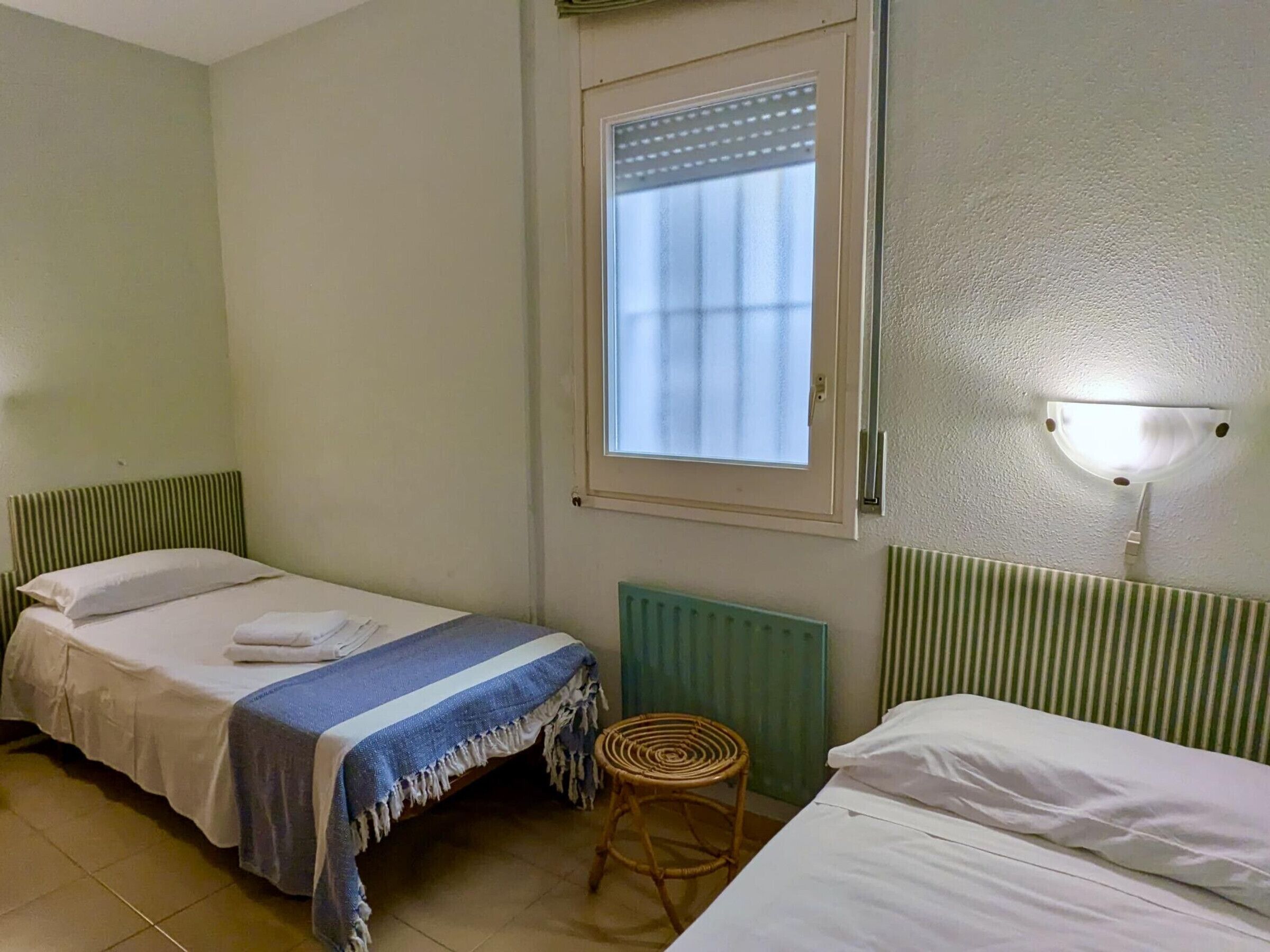 A small, simple room with two single beds, a window with blinds, a radiator, and a wall-mounted light fixture.