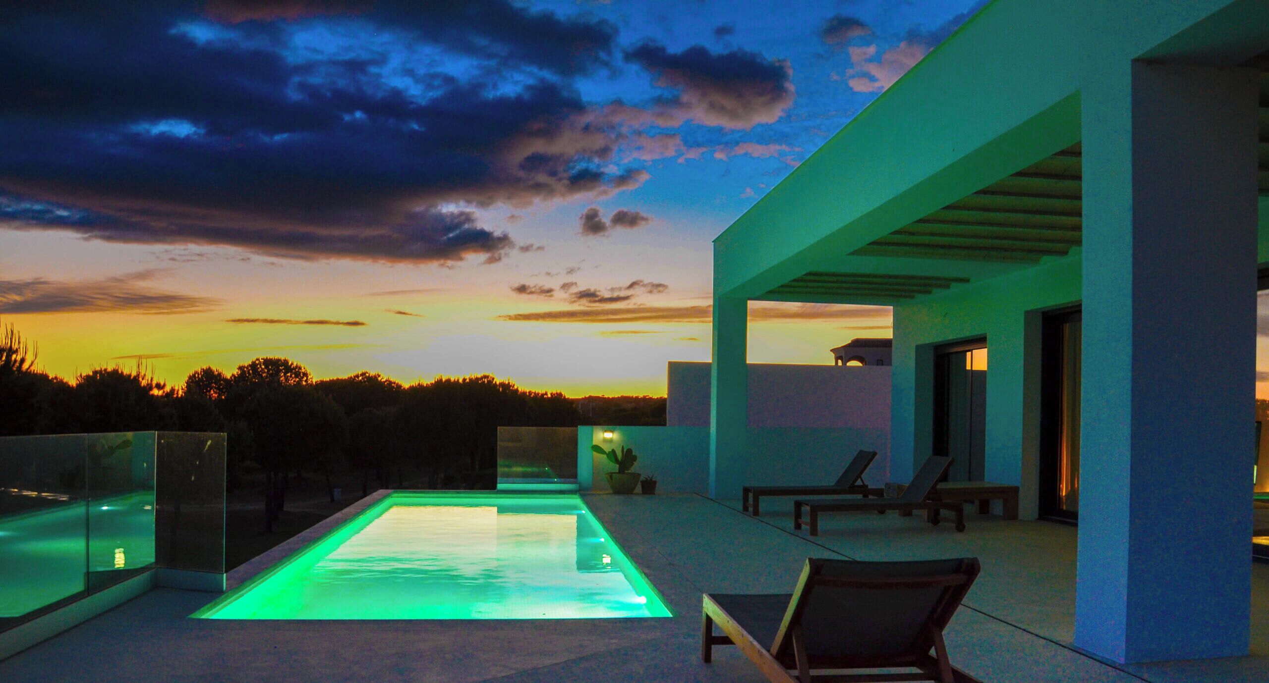 Un espace piscine extérieur moderne offrant une vue sur le coucher du soleil, avec des chaises longues et une balustrade en verre.