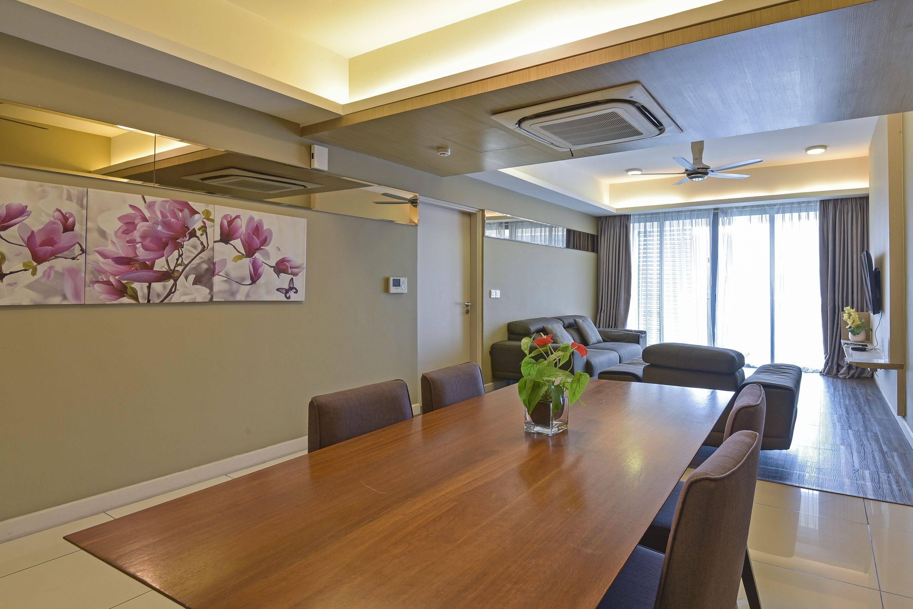A modern hotel room with a wooden dining table, comfortable seating, a large window with curtains, and a ceiling fan.