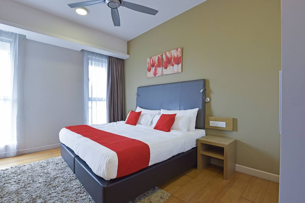 A neatly made bed with a red and white bedspread, a wooden nightstand, a ceiling fan, and a painting on the wall.