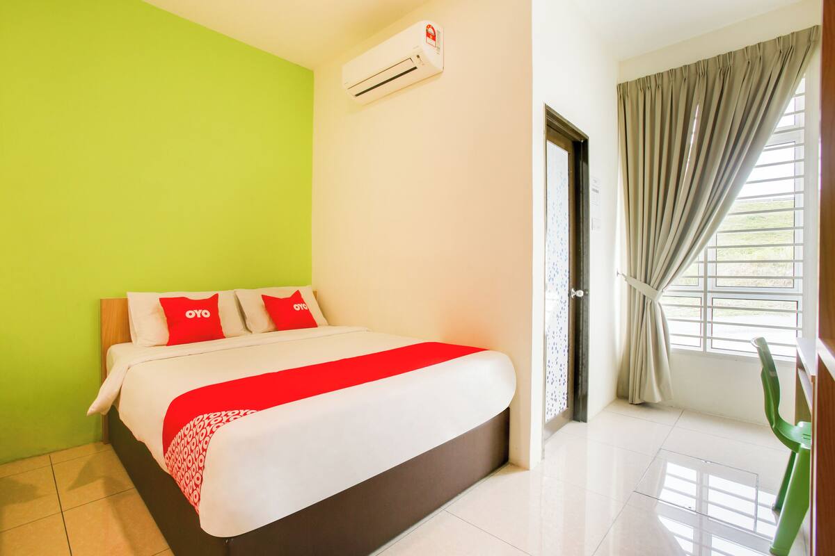 A hotel room with a bed, green walls, a window with curtains, and an air conditioning unit.
