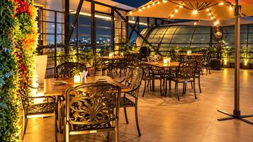 Un espace de restauration en plein air, avec des tables et des chaises, entouré de verdure et offrant une vue sur le paysage urbain la nuit.