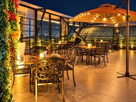 An outdoor dining area with tables and chairs, surrounded by greenery and a view of the cityscape at night.