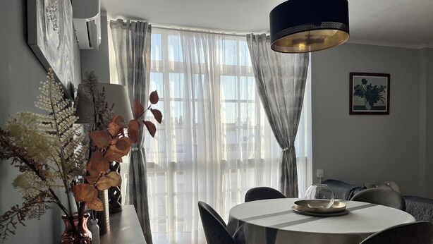 Spiseområde med et rundt bord, stole, en vase med tørrede blomster og en moderne pendel.
