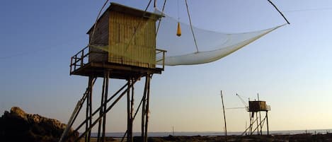 A beach with a wooden structure on stilts, a large white net, and a smaller wooden building in the background.