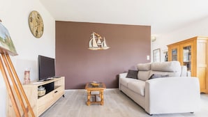 A living room with a beige sofa, a wooden coffee table, a television on a wooden stand, a wall clock, and a mural of a sailboat. - Quiet cottage near Pornic with wifi, pets welcome (Chauve)