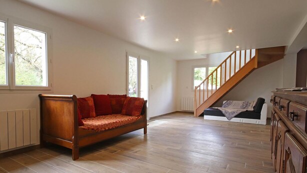Ein geräumiges Zimmer mit Holzboden, einer Holz-Couch, einer Treppe und Blick auf Bäume durch große Fenster.