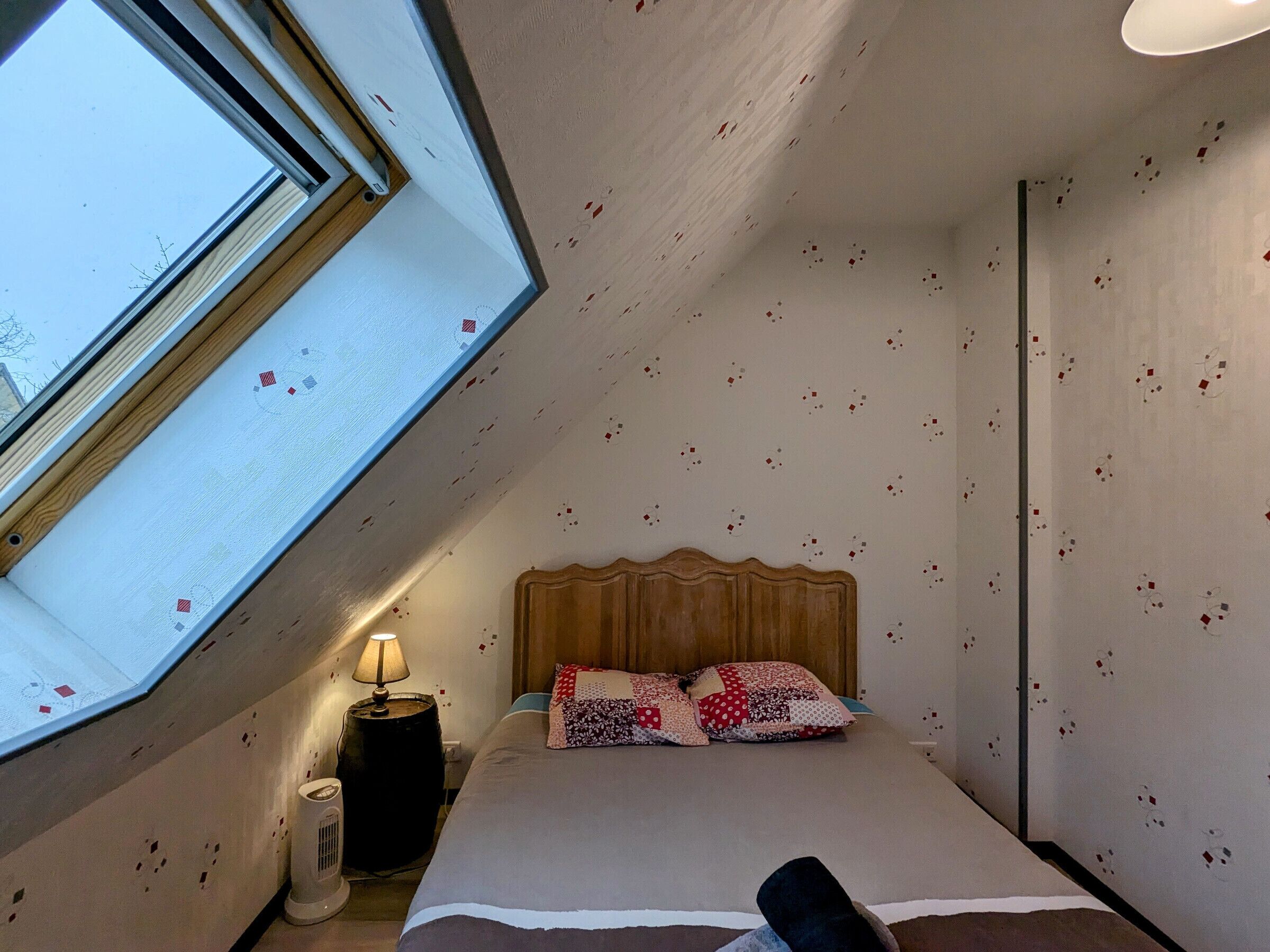 Un dormitorio con techo inclinado, cabecera de madera, cama con ropa de cama estampada, mesita de noche con lámpara y ventana con vista al cielo.