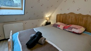 Ein Schlafzimmer mit schrägverdeckter Decke, einem hölzernen Kopfteil, einem Bett mit gemusterten Kissen, einer Nachttischlampe und einem Fenster mit Blick auf Bäume.