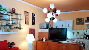 A compact living space with a sofa, a TV on a wooden cabinet, a small table with a lamp, and kitchen appliances in the background. - Cozy 1-room apartment with large terrace - in the middle of nature (Bad Harzburg)