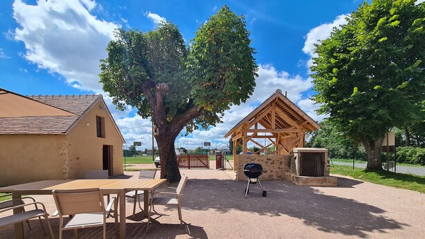 Un espacio al aire libre con mesa y sillas, una parrilla, un pabellón de madera y un árbol grande.