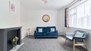 Un salon avec un canapé bleu, un fauteuil gris, une cheminée, un miroir et une fenêtre avec des rideaux.