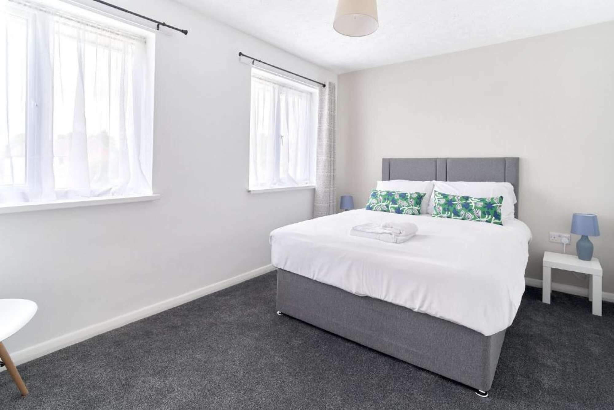A modern bedroom with a grey bed, white curtains, a grey headboard, and a small bedside table with a lamp.