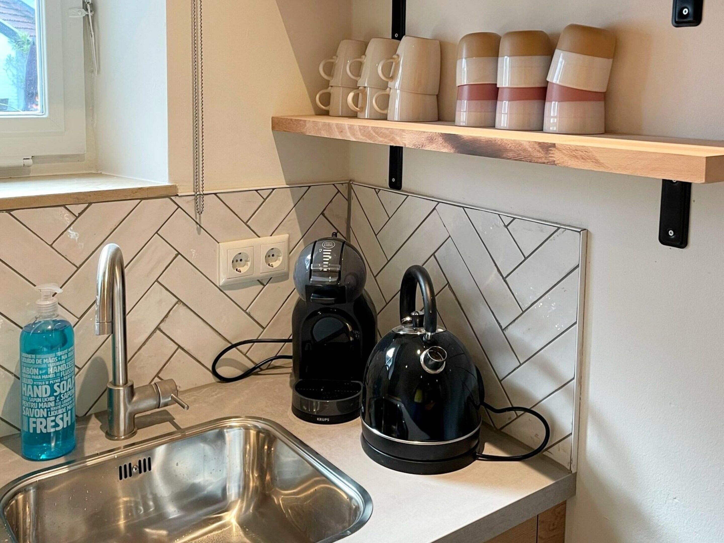 A kitchen with a sink, a kettle, a coffee maker, and a shelf with stacked cups.