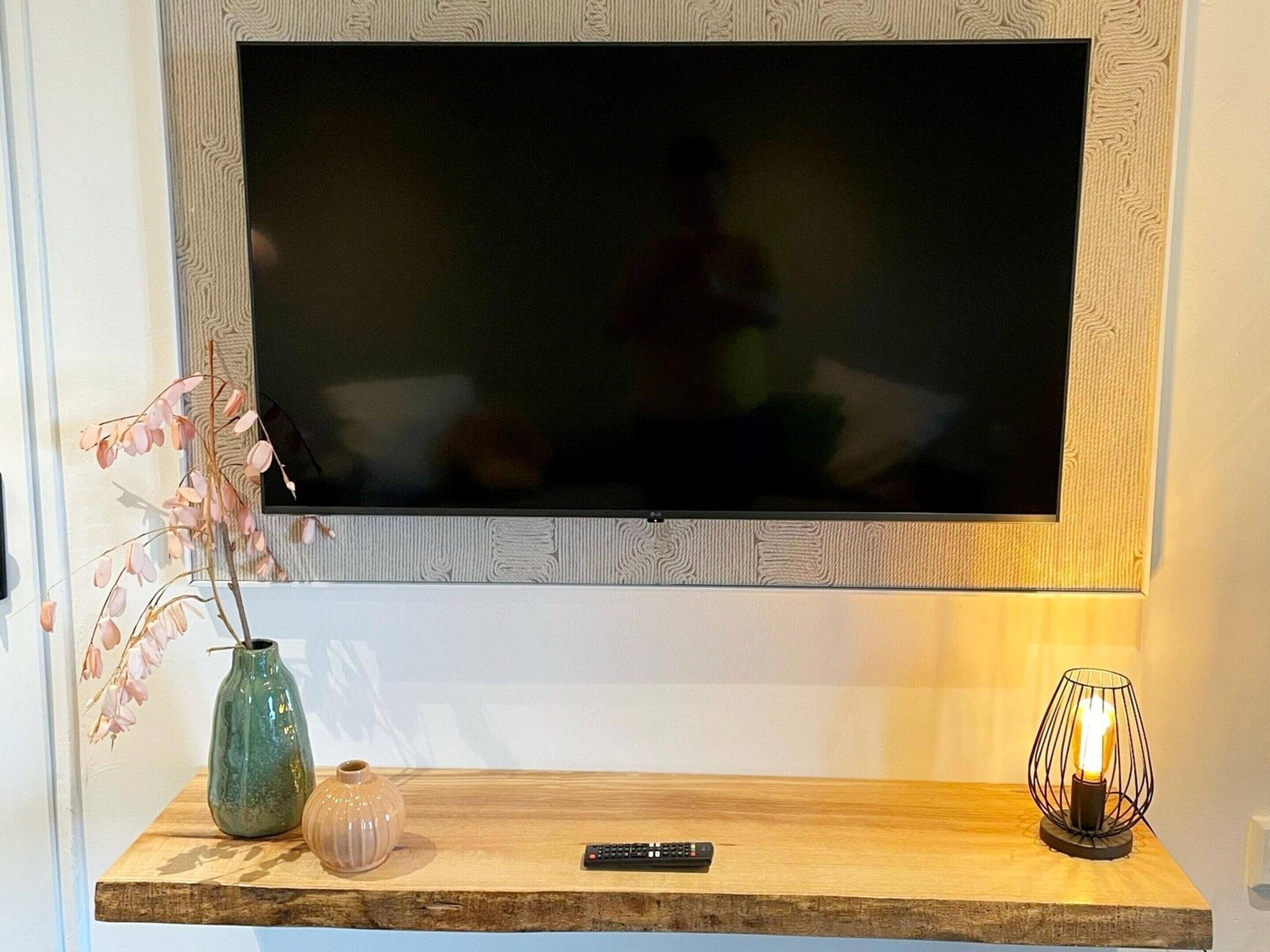 A flat-screen TV mounted on a wall with a wooden shelf below. On the shelf, there is a vase with flowers, a candle in a wire holder, and a remote control.