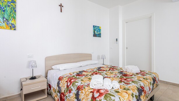 A neatly made bed with floral bedding, a bedside table with a lamp, and a wall-mounted cross. - Suore Figlie di San Francesco di Sales (La Spezia)