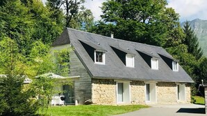 A two-story house with a stone facade and a sloped roof, surrounded by greenery and a paved driveway. - Mountain house in 24,000m² park. Pets allowed. (Aucun)