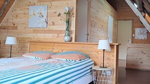 A cozy wooden cabin bedroom with a bed, two bedside lamps, a vase with flowers, and a door leading to another room. - Mountain house in 24,000m² park. Pets allowed. (Aucun)