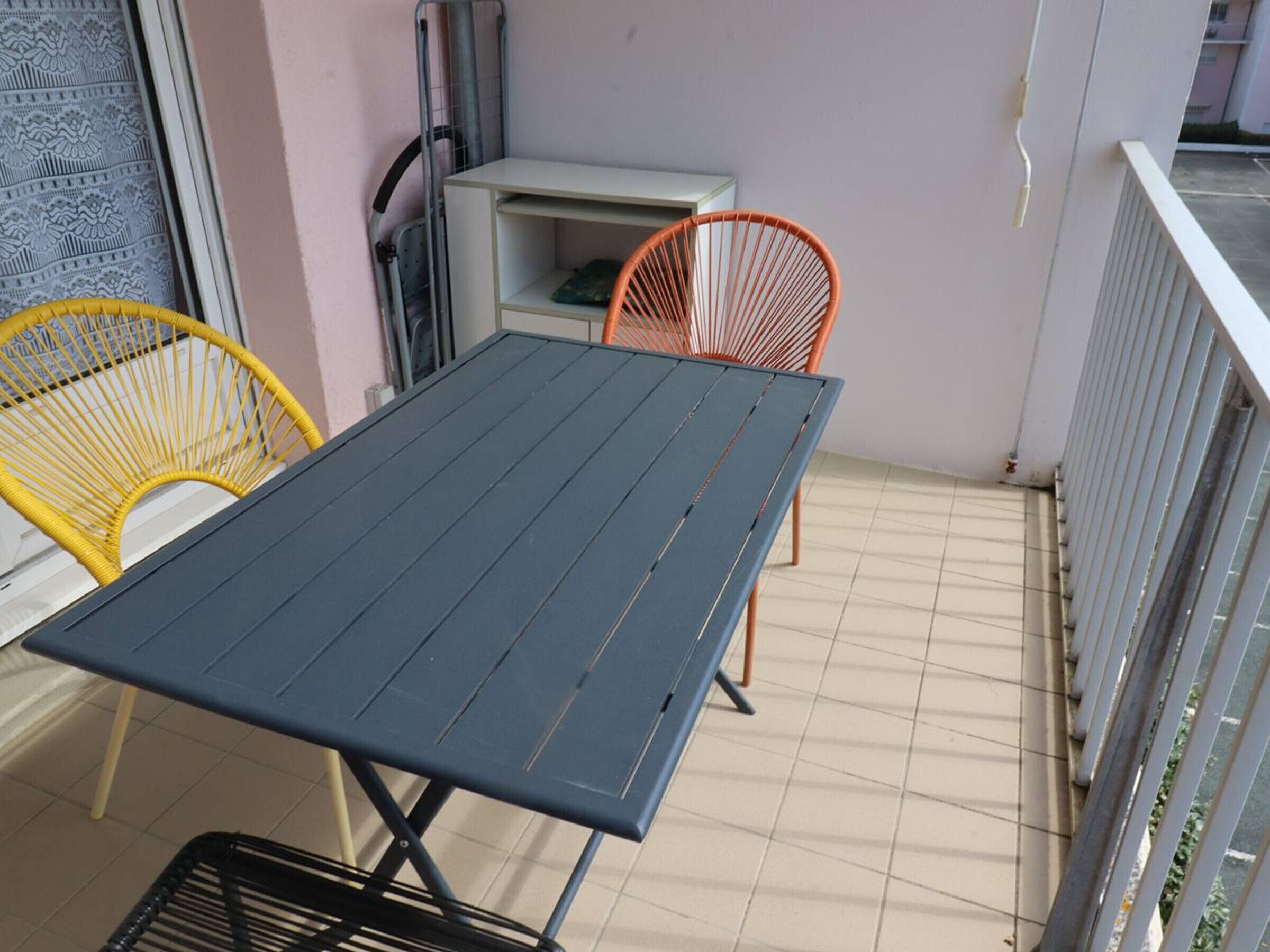 A balcony with a blue table and two chairs, one yellow and one orange.