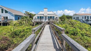 A wooden boardwalk leading to a beachfront house with a gabled roof and a covered porch.