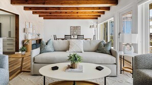 A modern living room with a sofa, coffee table, and side tables. There is a dining area in the background with a table and chairs.