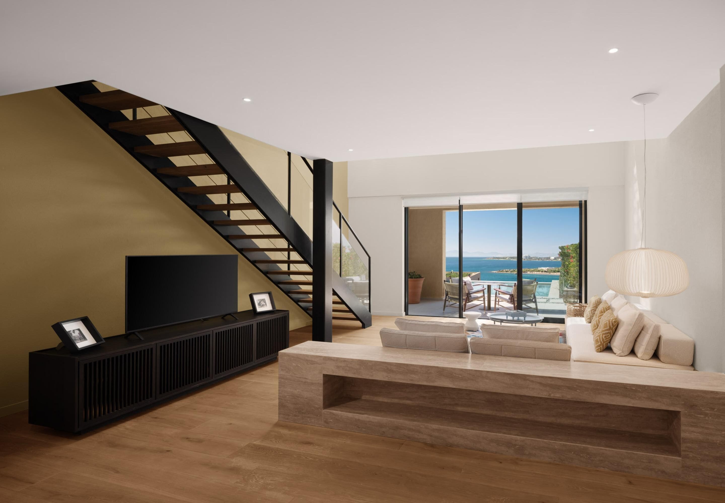 A modern living room with a staircase, a TV, a sofa, and a view of the sea.