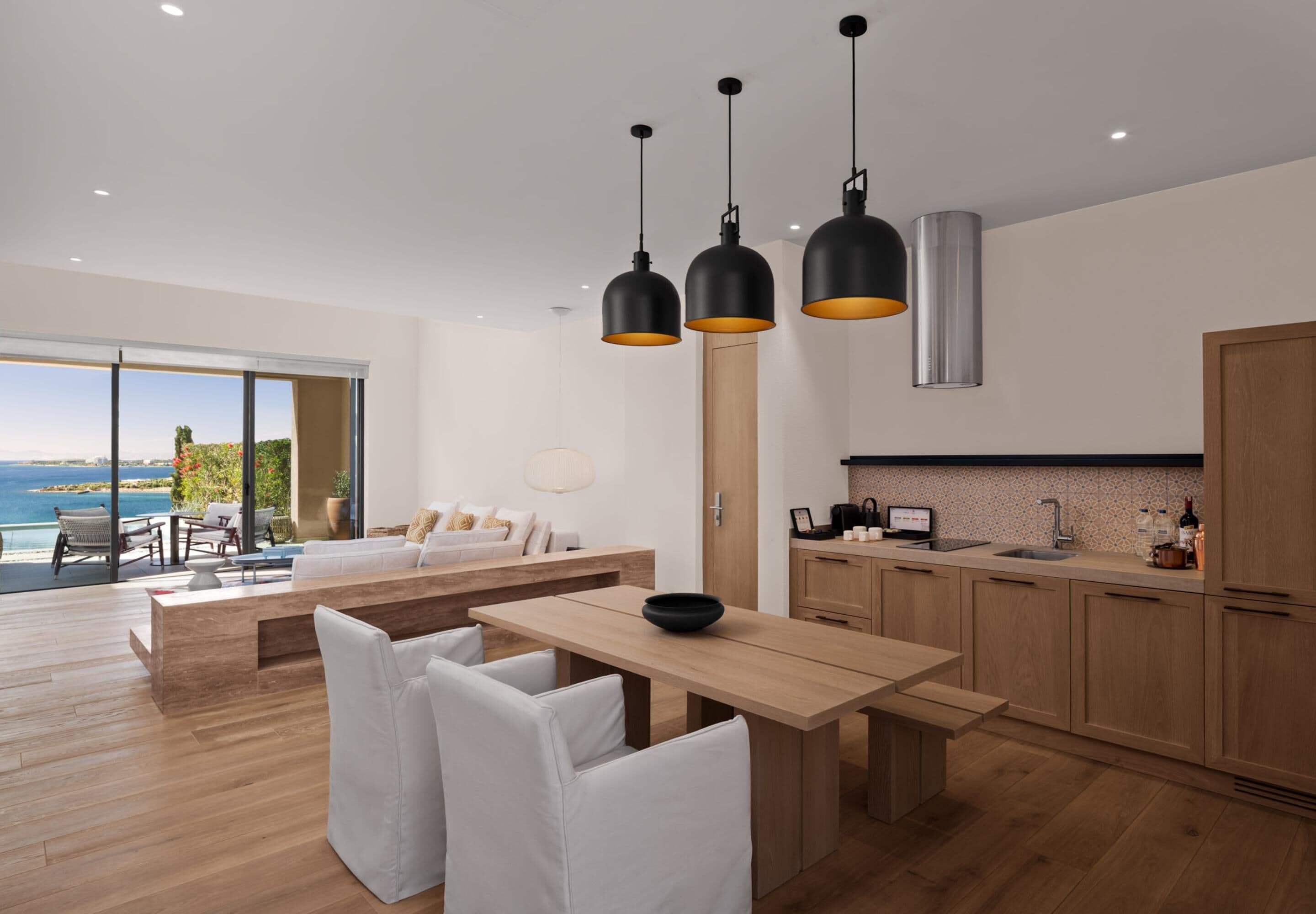 A modern kitchen with wooden cabinets, a dining area with white chairs, and pendant lights.