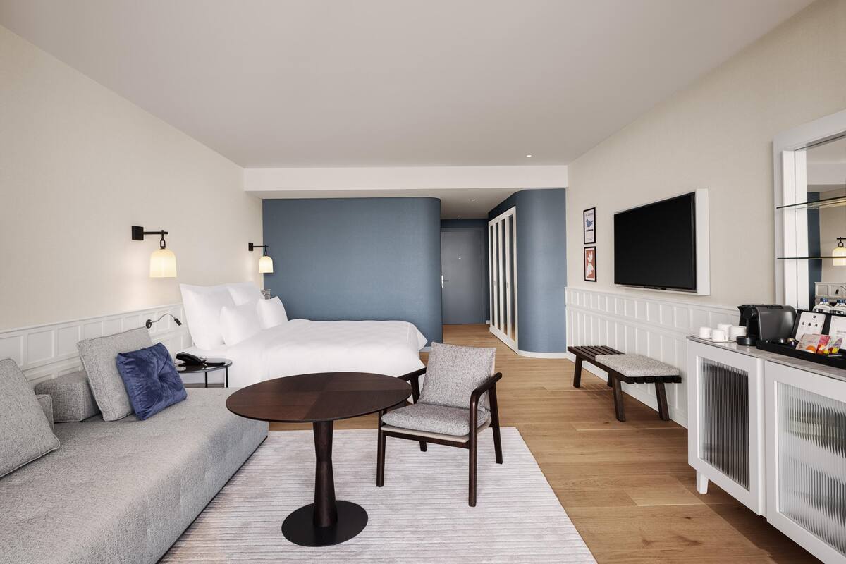 Una camera d'albergo con un letto, un divano, un tavolino, una panca, una televisione e un minibar.