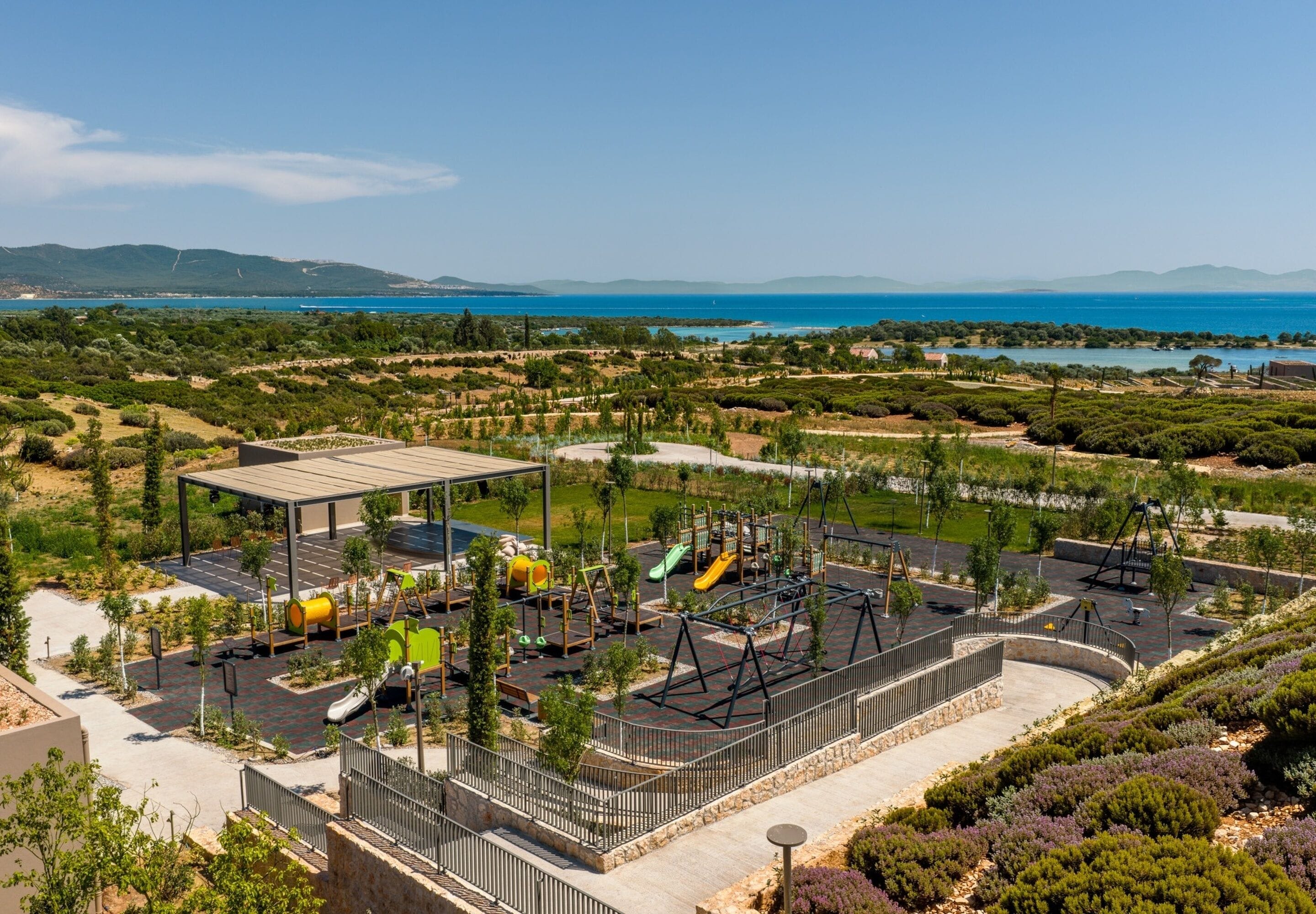 A playground with various play equipment, a covered seating area, and a view of the sea and hills in the background.